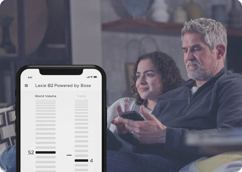Man watching TV with his daughter while adjusting his Lexie B2 hearing aids on the Lexie app.