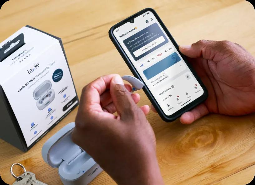 Person holding a smartphone with the Lexie app open, next to Lexie B2 Plus hearing aids and packaging on a wooden table.
