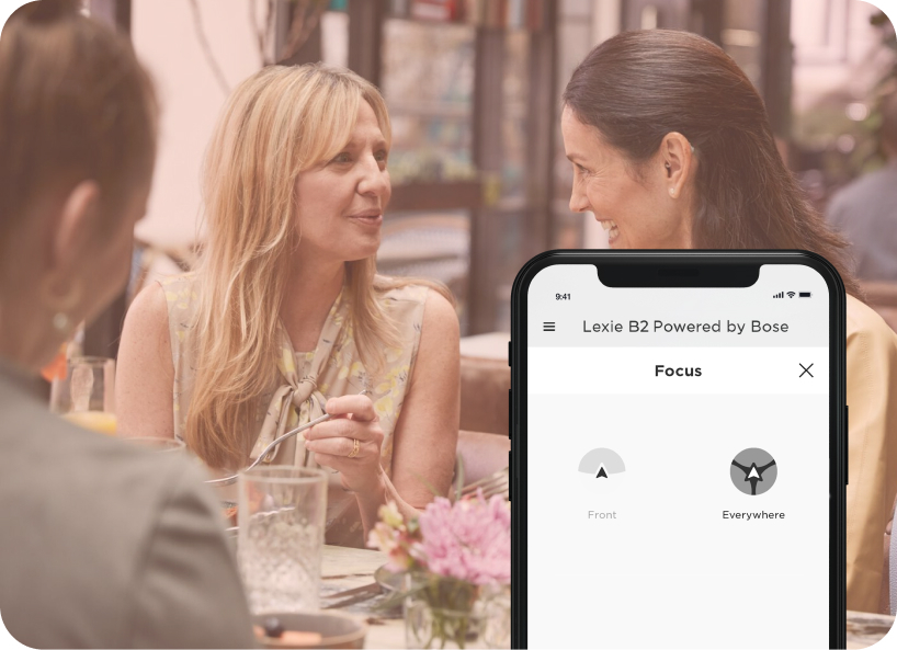 A group of woman eating at a restaurant, one adjusting her Lexie hearing aids on the Lexie app.
