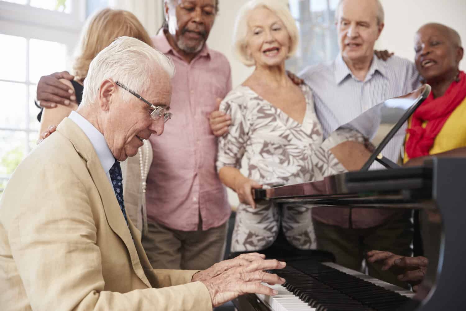 A group of 5 senior friends stand around the piano, playing and singing together after all of them took the most accurate online hearing test on the Lexie Hearing website to show the correlation between hearing loss and music