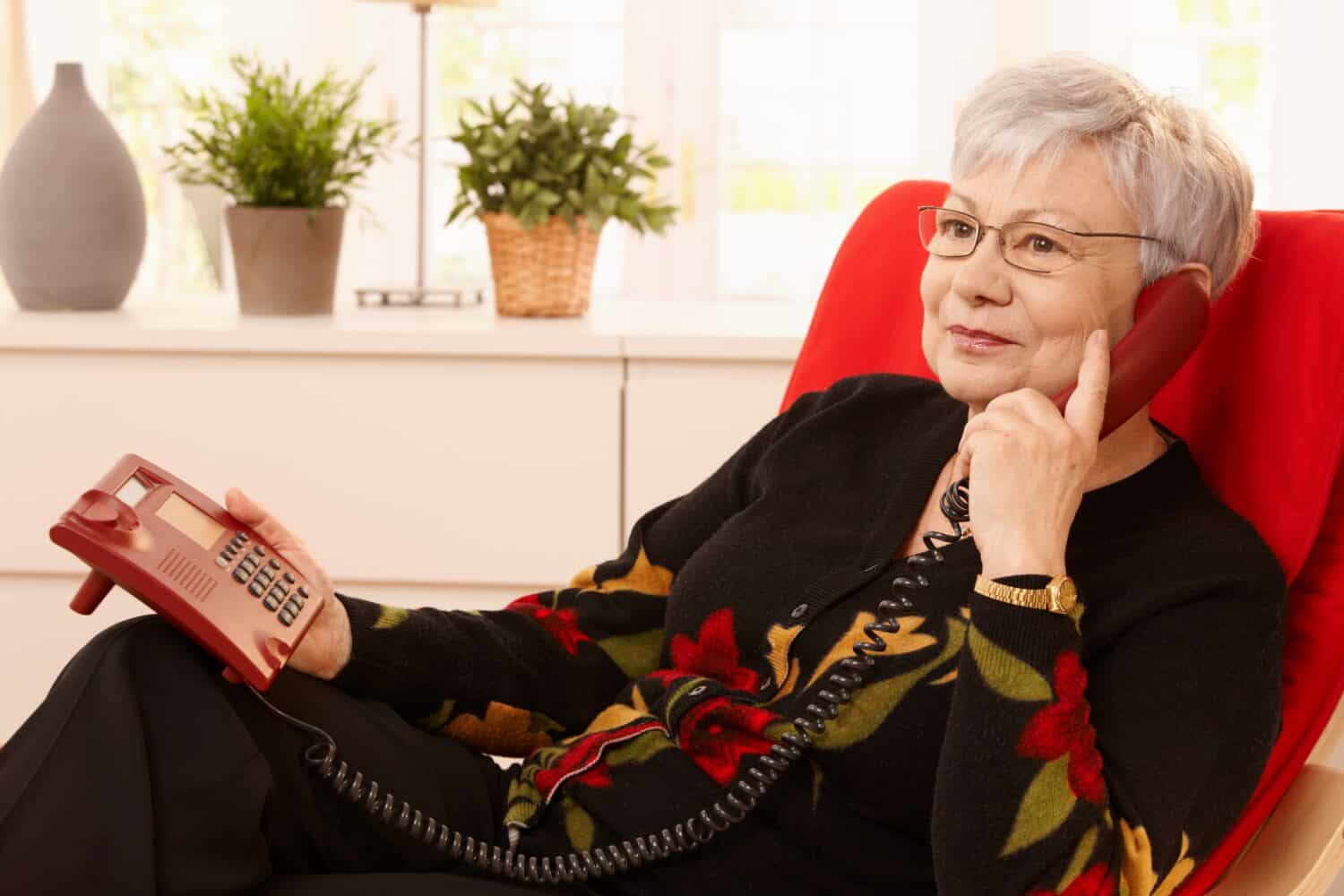 Senior woman sits on red chair talking on a landline telephone after she hears the phone ring through her best value hearing aid. can’t hear the phone ring
