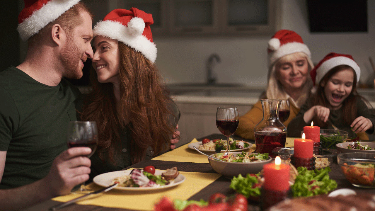 Hearing loss - Another year together. Happy male and female sitting at festive table and touching noses while drinking red wine. Senior lady with kid are on background. They are celebrating Christmas together