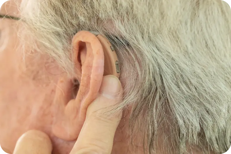 Behind shot of a senior man wearing a behind-the-ear beige hearing aid. He is adjusting the volume with his index finger on the hearing aid.
