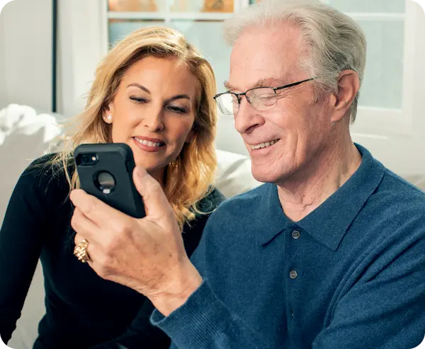 Senior man and middle-aged woman sitting on the couch. The man is holding a phone in his hand and they are both looking at the screen.
