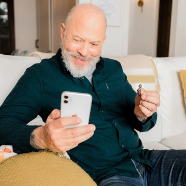 Senior man sitting on the couch holding a smartphone in one hand and a Lexie hearing aids in the other.